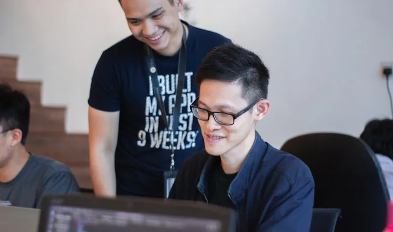 Two young professionals smiling and collaborating in front of a laptop, symbolizing mentorship and guidance.